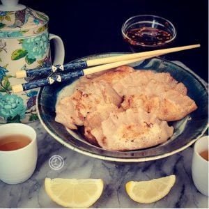 A bowl of potstickers, with lemon wedges, tea cups, teapot and chopsticks.
