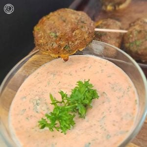 A bowl of Dairy-Free Harissa Dipping Sauce with a meatball on a toothpick over the sauce about to be dipped. With meatballs in the background and a sprig of parsley.