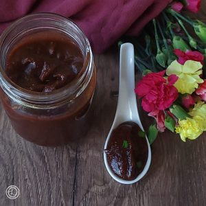 A jar and spoon full of the Hickory BBQ Sauce with a cloth napkin and flowers in the background.