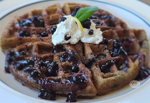 A closeup of a Huckleberry Waffle with Huckleberry sauce, a dollop of whipped cream, a sliver of lemon, and a sprig of mint.
