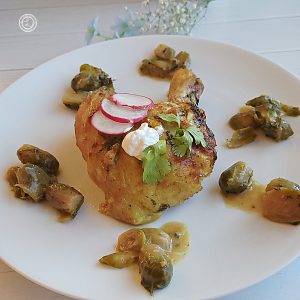 A square picture of the leg and thigh on a plate surounded by Brussel sprouts in a circular fashion, Topped with sliced radish, sour cream, and sprig of cilantro.