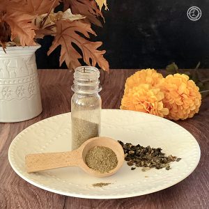 A plate with a small bottle of spice. A wooden spoon tilted against the bottle with spice. Raw dried spice on the plate, too. A vase with Autumn leaves and flowers in the background.
