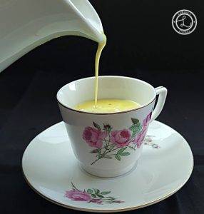 A white pitcher pouring latte into a flowered coffee cup and saucer.