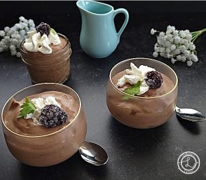 A close up of three dessert cups with whipped cream a blackberry and sprig of mint. Light blue pitcher and baby's breath in background.
