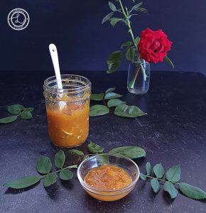 Apricot preserves in a jar with leaves and a rose in a vase in the background.