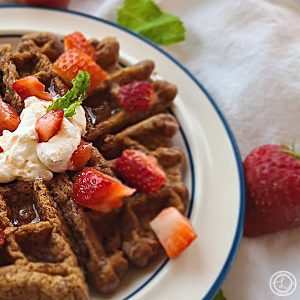 Gluten-Free Fresh Strawberry Waffles a quarter of a waffle poking out the left side. Topped with chopped strawberries, whipped coconut milk, and sprig of mint.