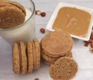 Gluten-Free Peanut Butter Sandwich Cookies pictured is a cookie being dunked in milk with other cookies and peanut butter in a small bowl and peanuts to the side of the bowl.