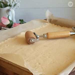 A square picture with flowers and a pitcher in the background. Gluten-Free Family Size Pizza Crust in the center of the photo with a rolling pin in the center.