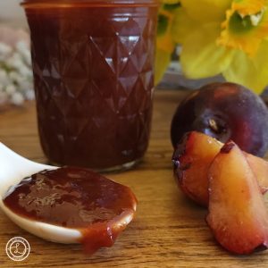 Small Batch Plum Preserves in a jar and some on a spoon with a plum, plum slices, and flowers in the background.
