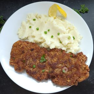 A large Country Fried Pork Chop on a plate wit mashed potatoes and chives.