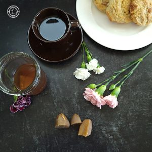 Stem Ginger in syrup with three ginger pieces in front. A jar with syrup, carnations, coffee cup, and saucer with scones in background.