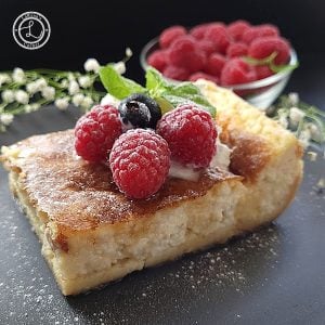 Gluten-Free Blintz Casserole with whipped coconut cream, raspberries, blueberries, and sprig of mint. In the background is baby breath flowers and a bowl of raspberries.