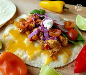 A chicken taco on a cutting board with tortillas on the side with lime slices and baby bell peppers on surrounding it.