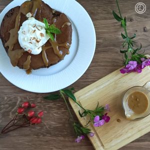 Peanut Butter Glaze in a pitcher. A plate of chocolate pancakes with the glaze whipped coconut milk, and chopped peanuts, with sprig of mint and flowers.