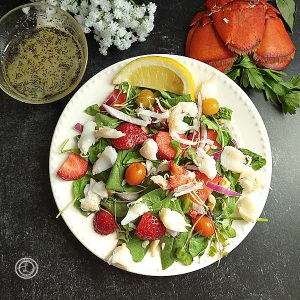 Spinach Strawberry Lobster Salad on a plate with a little of a lobster tail in the background and flowers. Dressing off to the side in a bowl.