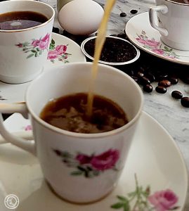 A cup of Swedish Egg Coffee being poured with two cups, and egg, and coffee beans in the background.