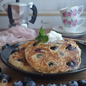 Gluten-Free Blueberry Cinnamon Pancakes on a plate with whipped coconut cream and a sprig of mint. An espresso maker and espresso cups in the background.