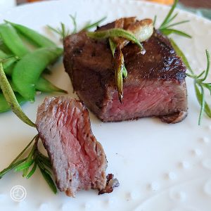 An Elk Steak with a cut into with a piece on the side. With sugar snap peas, rosemary sprig, and roasted garlic on top of the steak.
