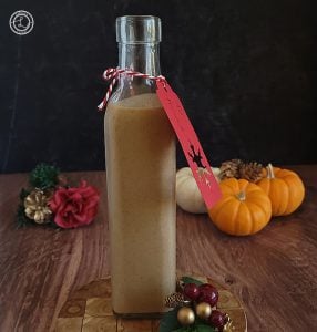 Homemade Pumpkin Cream Liqueur in a bottle with a gift card tied on. In the background is a pinecone with flower and leaves and three mini pumpkins with small pinecones on top of them. A pinecone next to the bottle.