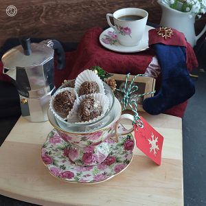 A coffee cup and saucer with Swedish Chocolate Balls inside, and espresso maker, a espresso teacup and saucer, a box of chocolates, and a small Christmas stocking.