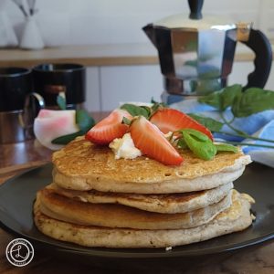 Gluten-Free Buttermilk Pancakes topped with whipped cream, mint leaves, and fresh cut strawberries. Flower, espresso cup, and espresso carafe in the background.