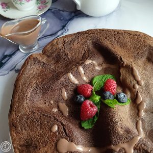Gluten-Free Mocha Sauce on a Chocolate Dutch Pancake with a small gravy boat of the sauce in the background. Berries and sprig of mint on top of pancake.