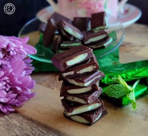 A stack of Homemade After-Dinner Chocolate Mints in front of a bowl of mints. Three mints wrapped in green foil wrappers.