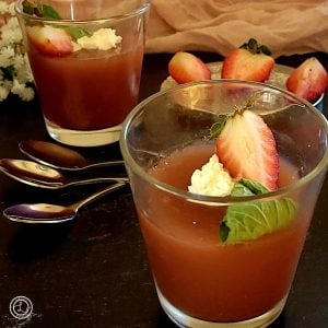 Two Servings of Homemade Strawberry Jello with spoons, whipped coconut cream, strawberry wedge, and sprig of mint. Strawberries in the background.