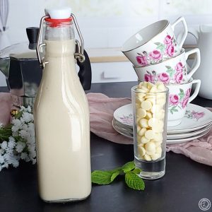 Close up of a bottle of Homemade White Chocolate Syrup and a shot glass of white chocolate chips.