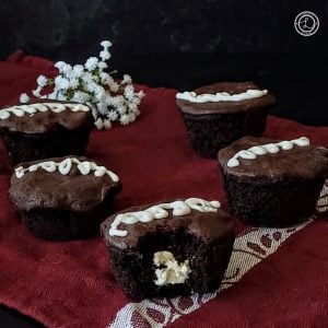 Three Gluten-Free Hostess Cupcakes on a tablecloth with flowers in the background. Center cupcake has a bite out of it. You can see the filling.