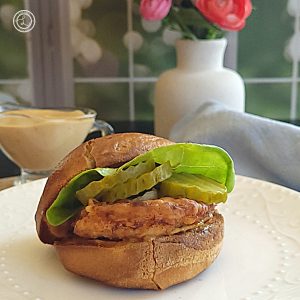 Gluten-Free Copycat Chick-Fil-A Sandwich a close up of the sandwich with lettuce and pickles. Behind it is a small container of sauce, a vase with flowers, and a napkin.