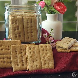 Gluten-Free Graham Crackers in a glass jar with crackers in front and to the side. A vase with flowers behind it.