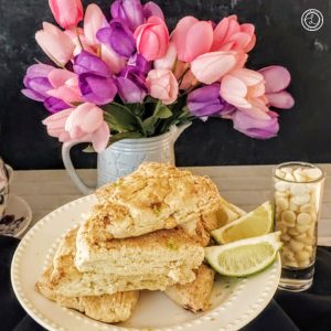 Gluten-Free Lime White Chocolate Scones in a stack on a plate with lime wedges. A small shot glass with white chocolate chips and flowers in a vase in the background.