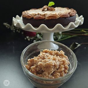 Dairy-Free Pecan Coconut Frosting a bowl of frosting in front of a cake topped with frosting, sliced cherries, and a sprig of mint.