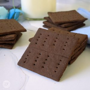 Gluten-Free Chocolate Graham Crackers three stacks in front of a large glass of milk and flowers to the side.