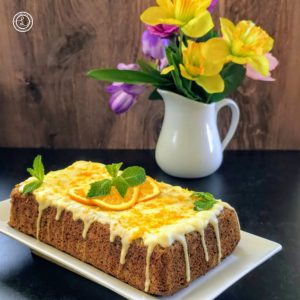 Gluten-Free Orange Banana Bread on a platrer topped with orange glaze, orange zest, 3 orange slices, and 3 sprigs of mint. A vase of flowers in the background.