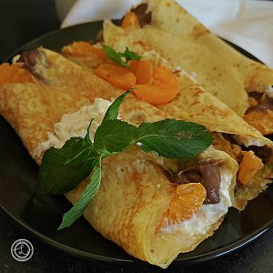 Gluten-Free Sweet Dessert Crepes filled with vegan chocolate pudding, coconut whipped cream, and orange segments. Topped with whipped coconut cream, madarine segments, and sprig of mint.