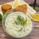 The Best Tartar Sauce in a bowl in front of a plate of beer battered fish. The sauce is topped with dill sprig and lemon twisted lemon slice.