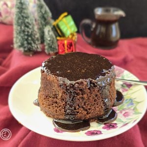 Gluten-Free Sticky Toffee Pudding. Sticky toffee pudding on a small plate on top of a red table cloth. Two small two pine tree, two small wrapped gifts. Sicky Toffee Sauce in a small pitcher in the background.