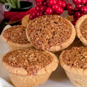 Gluten-Free Pecan Tassies. A platter of tassies in a square photo with berries in the background.